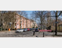 Kreuzung in einem Wohngebiet mit mehrstöckigen, rosafarbenen Häusern auf der linken Seite, kahlen Bäumen entlang der Straße und parkenden Autos. Im Vordergrund sind Absperrungen und Straßenschilder sichtbar, darunter ein Schild mit einer Geschwindigkeitsbegrenzung von 30 km/h.