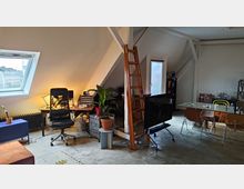 Ein lichtdurchfluteter Raum mit Dachschräge, eingerichtet als kombiniertes Büro und Wohnbereich. Sichtbar sind ein Schreibtisch mit einem Bürostuhl, Pflanzen, eine hölzerne Leiter, ein großer Esstisch mit verschiedenen Stühlen sowie Dachfenster und weiße Balkenkonstruktionen.