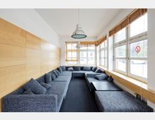 L-förmiger Aufenthaltsraum mit grauem Ecksofa und zahlreichen Kissen. Die Wände bestehen aus Holzpaneelen und großen Fenstern mit Jalousien, durch die Tageslicht in den Raum fällt; zwei Hängeleuchten hängen von der Decke.