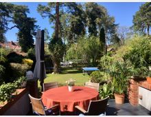 Garten mit Terrasse, auf der ein runder Tisch mit roter Tischdecke und Stühlen steht. Im Hintergrund sind grüne Rasenflächen, Bäume, ein Tischtennistisch und mehrere Sträucher sowie ein kleiner Pavillon zu sehen, umgeben von Wohnhäusern mit roten Dächern.