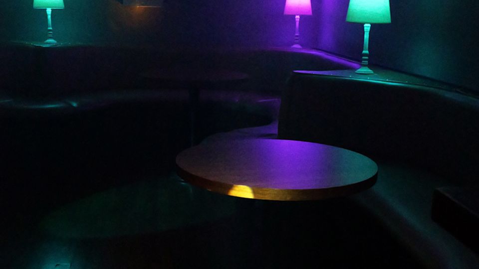 Dunkler Lounge- oder Clubbereich mit halbrunden Polsterbänken entlang der Wände und einem runden Tisch in der Mitte. Bunte Akzentbeleuchtung in Türkis und Violett sowie kleine Tischlampen und Lichtpunkte auf dem Holzboden prägen die Szene.