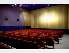 Kinosaal mit Reihen von orangefarbenen Polstersitzen, einer großen Bühne mit geschlossenem gelben Vorhang und dunkelblauen Wänden. An der Decke befinden sich kleine Leuchten, die den Raum beleuchten.