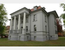 Großes, zweistöckiges Gebäude im klassizistischen Stil mit weißer Fassade, vier großen Säulen an der Vorderseite und mehreren hohen Fenstern mit Rundbögen. Das Haus steht inmitten einer Grünfläche und ist von Bäumen umgeben.