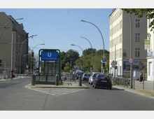 Straßenszene mit einer U-Bahn-Station der Linie U6, Haltestelle „Schwartzkopffstraße“. Im Vordergrund eine gläserne Eingangskonstruktion, umgeben von geparkten Fahrrädern, Autos und Fußgängerwegen; links und rechts stehen mehrstöckige Gebäude, im Hintergrund sind Bäume sichtbar.