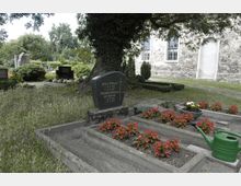 Ein kleiner Friedhof mit mehreren Gräbern, die mit roten Blumen bepflanzt sind und auf einem von Bäumen beschatteten Gelände liegen. Im Hintergrund ist ein Gebäude aus Natursteinmauerwerk mit hohen Fenstern zu sehen.