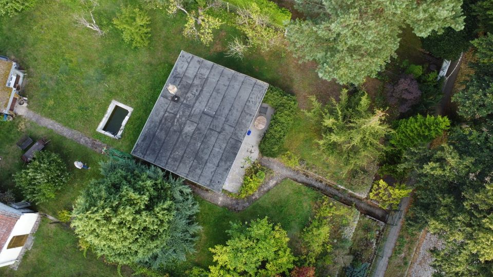Luftaufnahme eines Grundstücks mit einem rechteckigen Gebäude mit flachem, dunklem Dach, umgeben von Rasenflächen und dichtem Baumbestand. Sichtbar sind ein kleiner rechteckiger Wasserbehälter oder Teich, eine Terrasse am Haus sowie ein schmaler Weg, der zu einem Tor am Grundstücksrand führt.