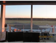 Blick aus einem Flughafen-Tower mit großen Fenstern auf Rollfelder, Grünflächen und eine Baumreihe im Hintergrund. Im Vordergrund sind technische Kontrollgeräte und Bildschirme sichtbar.