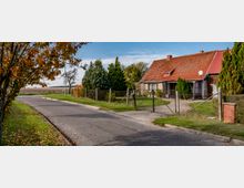 Ländliche Straße mit einem freistehenden Wohnhaus, das ein rotes Ziegeldach und mehrere kleine Fenster hat. Das Grundstück ist von einem Maschendrahtzaun umgeben, und im Vordergrund sind Bäume mit herbstlich gefärbtem Laub sowie ein asphaltierter Gehweg zu sehen.