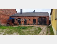 Backsteinbau mit grauem Dach und großen Holztoren auf einem Hof mit gepflastertem und bewachsenem Boden. Links stehen Gartenmöbel und ein Grill unter einem blauen Sonnensegel; rechts ist eine gelb gestrichene Hauswand zu sehen.