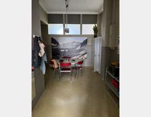 Innenraum eines schlichten Aufenthalts- oder Pausenraums mit rundem Tisch und roten Stühlen vor einer großen Fotowand mit Berglandschaft. Hohe Fenster mit Rollos, graue Wände, ein weißer Spind und ein Servierwagen mit Küchenutensilien prägen den Raum.