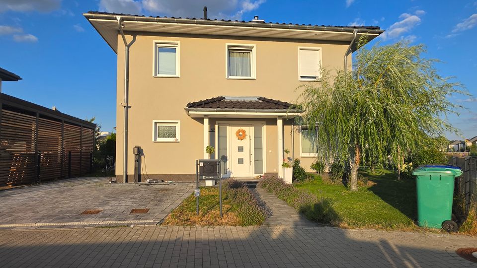 Zweistöckiges, beige verputztes Einfamilienhaus mit dunklem Satteldach und kleiner überdachter Eingangsnische mit weißer Haustür. Vorn liegen eine gepflasterte Einfahrt, ein schmaler Gartenweg mit Beeten und ein kleiner Rasen mit Baum; rechts steht eine grüne Mülltonne am Straßenrand.