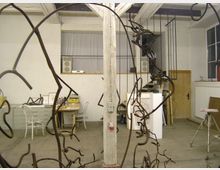 Ein Künstleratelier mit weißen Wänden, Holzbalken an der Decke und einem Betonboden. Im Vordergrund sind metallische Skulpturen aus gebogenen Stäben, im Hintergrund Arbeitsflächen mit Stühlen, Schränken und Materialien sowie eine Holztreppe zu sehen.