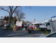 Straßenmarkt mit Verkaufsständen und Lieferwagen in einer gepflasterten Straße. Im Vordergrund sind Tische mit verschiedenen Waren, im Hintergrund ein Transporter mit der Aufschrift "Landfleischerei" und eine Uhr auf einem Mast.