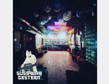 Ein stimmungsvoll beleuchteter Nachtclub mit einer zentralen Bar im Hintergrund, umgeben von gepolsterten Sitzgelegenheiten und dekorativen Wänden. Die Decke ist mit schimmernden Discokugeln und bunten Lichtern gestaltet.