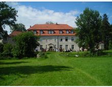 Herrenhaus mit heller Fassade und rotem Ziegeldach, umgeben von einer großzügigen Grünfläche mit Bäumen; Menschen sind vor dem Gebäude versammelt, und der Himmel ist teils bewölkt.