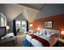 Helles Hotelzimmer mit einem Doppelbett, orangefarbener Akzentwand und großen Fenstern, die zu einem Balkon mit Blick auf einen Leuchtturm und eine Wasserlandschaft führen. Das Zimmer verfügt über moderne Möbel, darunter einen Fernseher, zwei Nachttische und eine gemütliche Sitzecke.