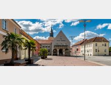 Marktplatz einer kleinen Stadt mit dem Rathaus von Trebbin im Zentrum, erkennbar an der Fassade mit Arkaden und einem kleinen Turm. Rundherum befinden sich bunte, historische Wohnhäuser und dekorative Palmen in Pflanzkästen, bei klarem Himmel und Sonnenschein.