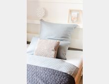 Ein einfaches Bett mit gestreifter hellblauer Bettwäsche, einem beigen Zierkissen und einer dunkelgrauen Steppdecke. Im Hintergrund sind eine weiße Holzwand, ein dekorativer Ornamentanhänger und ein gerahmtes Bild zu sehen.