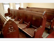 Reihen von dunkelbraunen, holzgeschnitzten Kirchenbänken mit dekorativen Mustern, teilweise beleuchtet durch Fenster mit Gitterverglasung im Hintergrund. Der Boden ist teilweise mit einem roten Teppich bedeckt.