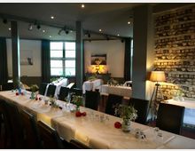 Ein Veranstaltungsraum mit gedeckten Tischen, dekoriert mit weißen Tischdecken, Blumen, Gläsern und kleinen roten Akzenten. Im Hintergrund sind eine Steinwand, ein großes Fenster und eine Stehlampe zu sehen.