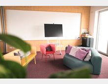 Ein modern gestalteter Besprechungsraum mit bunten Polstermöbeln auf einem weinroten Teppichboden, einer großen weißen Tafel an der Wand und einem mobilen Bildschirm auf einem Ständer. Im Hintergrund befinden sich ein Regal mit Büromaterialien und eine großzügige Fensterfront, die Tageslicht hereinlässt.