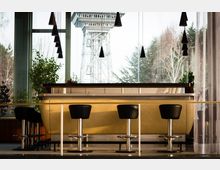 Innenaufnahme einer modernen Bar mit gepolstertem gelbem Tresen und schwarzen Barhockern, umgeben von großen Glasfenstern. Im Hintergrund ist ein Außenausblick mit Bäumen und einem Stahlgerüst-Turm sichtbar, begleitet von hängenden, kegelförmigen Lampen.