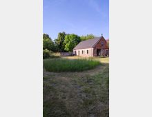 Ein Backsteinhaus mit Schrägdach in ländlicher Umgebung, umgeben von einer Wiese mit kurzem und hohem Gras. Im Hintergrund sind Bäume und ein hölzerner Zaun zu sehen, der den Hof abgrenzt.