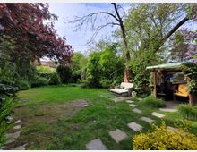 Grüne Gartenlandschaft mit zentraler Rasenfläche, umgeben von Bäumen, Sträuchern und buntem Blattwerk. Rechts sind eine überdachte Sitzecke mit Sofa und ein kleiner Holzschuppen sichtbar, durchzogen von einem gepflasterten Weg.