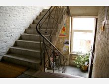 Treppenhaus mit groben gemauerten Wänden, einem Geländer aus Metall und Betonstufen. Im Hintergrund befindet sich ein großes Fenster, durch das Tageslicht einfällt, sowie eine Topfpflanze und Poster an den Wänden.