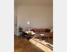 Wohnzimmer mit hellen Holzdielen, einem großen rosafarbenen Sofa, einem schwarzen Sessel mit Metallrahmen und einem kleinen Glastisch. Eine grüne Zimmerpflanze steht auf einem Pflanzenständer, und ein großer Teppich in warmen Orangetönen bedeckt den Boden.