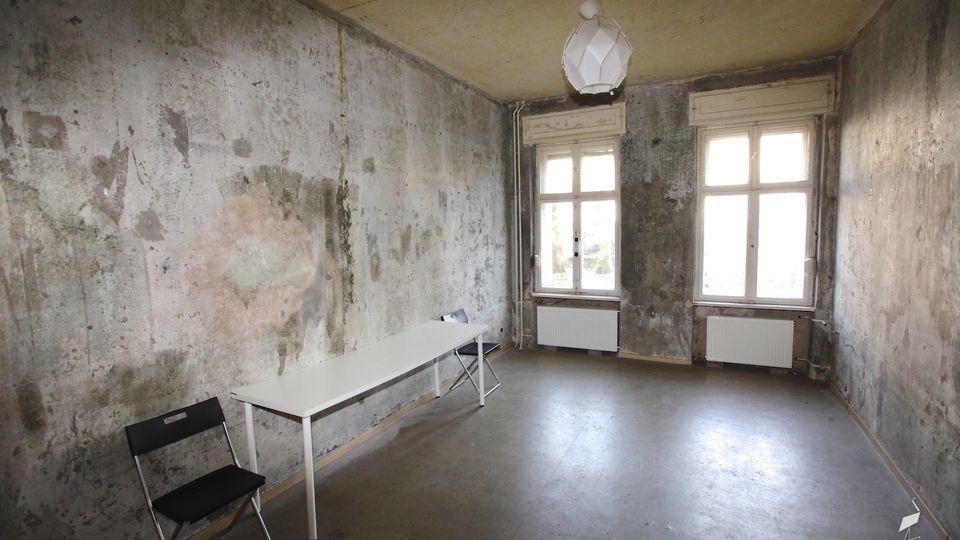 Karger, leerer Innenraum mit abgenutzten, fleckigen Putzwänden und blankem Boden, spärlich möbliert mit einem langen Tisch und Klappstühlen. An der Stirnseite befinden sich zwei hohe Fenster mit Heizkörpern darunter und eine einfache Deckenleuchte.