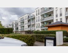 Moderne Wohnanlage mit weißen, mehrstöckigen Gebäuden, Balkonen und großen Fenstern. Im Vordergrund befinden sich gepflegte Hecken, ein Holzschuppen und eine Beschilderung mit der Adresse "Elisabeth-Feller-Weg 4-10".