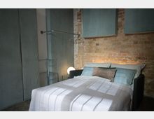 Ein Schlafzimmer mit einem Bett, das weiße Bettwäsche und mehrere Kissen in Blau- und Brauntönen hat. Die Wände bestehen aus freiliegenden Ziegelsteinen und sind teilweise mit Stoffplatten verkleidet; daneben steht ein Metallregal mit einer leuchtenden Kugellampe.