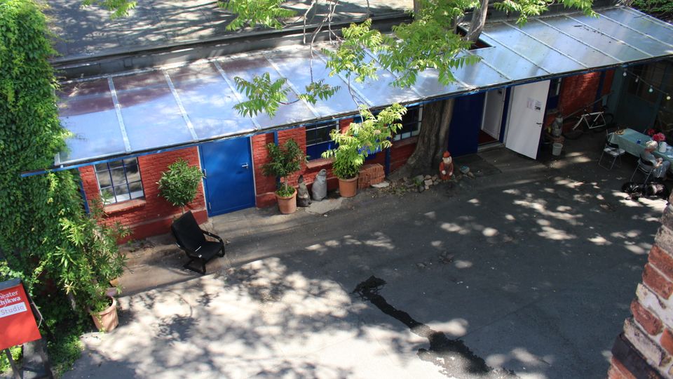 Innenhof mit einem niedrigen, langgezogenen Backsteingebäude, blau gestrichenen Türen und Fenstern sowie einem flachen, teils transparenten Dach. Vor dem Gebäude stehen Topfpflanzen; Bäume werfen Schatten auf den Asphalt, daneben sind ein Fahrrad und ein kleiner Sitzbereich zu sehen.