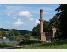 Backsteinbau mit einem hohen, runden Turm und Zinnen, gelegen an einem Fluss mit grüner Uferlandschaft und Bäumen im Hintergrund. Links im Bild befindet sich ein kleines Boot auf dem Wasser, daneben ein Gebäude hinter dichter Vegetation.