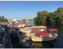 Ein Bootsanleger an einem Fluss mit mehreren kleinen Motorbooten, teils mit roten und beigen Abdeckungen. Im Hintergrund sind grüne Bäume, Gebäude sowie ein blauer Himmel sichtbar.