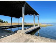 Überdachter Bootsanleger mit Betonpfeilern und Holzstegen, umgeben von Schilf am Ufer eines ruhigen Sees unter klarem blauem Himmel.