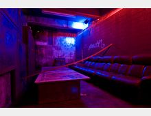 Ein dunkler Raum mit Betonwänden, beleuchtet von blauen und roten Neonlichtern. Im Vordergrund steht ein niedriger Tisch, dahinter eine lange, schwarze Ledercouch und Wände mit Graffiti.