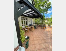 Terrasse eines Hauses mit rotbraunen Fliesen und einer schwarzen Holzkonstruktion für einen Überbau. Darunter befinden sich ein Holztisch, Rattan- und Gartenstühle sowie Pflanzen und Gartenutensilien. Im Hintergrund sind Bäume und umliegende Gebäude sichtbar.