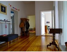 Ein heller Flur in einer Wohnung mit Holzdielenboden, mehreren weißen Türen und minimaler Dekoration, darunter Kleider auf einem Holzständer, ein kleines schwarzes Sofa und ein antiker Holztisch. Im Hintergrund sind durch offene Türen weitere Räume mit Möbeln und Pflanzen sichtbar.