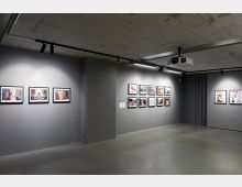 Ein moderner Ausstellungsraum mit grauen Wänden, an denen gerahmte Fotografien in gleichmäßigen Abständen hängen. Die Decke besteht aus unbehandeltem Beton, und die Beleuchtung erfolgt über Schienenstrahler.