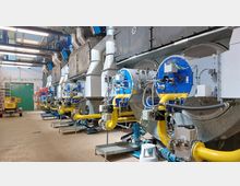 Industrielle Anlage mit mehreren großen Kesseln und Rohrleitungen in einer Halle. Die Kessel sind mit blauen und gelben Komponenten ausgestattet, umgeben von Stahlrohren und moderner Maschinentechnik.