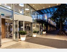 Eingangsbereich eines modernen Gebäudes mit rotierender Glastür, großen Fenstern und beleuchteten Pflanzkübeln. Im Hintergrund eine ruhige Straßenansicht mit Bäumen, geparkten Fahrrädern und Autos.