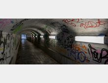Ein schmaler, gewölbter Fußgängertunnel mit weiß gekachelten Wänden, die mit bunten Graffiti bedeckt sind. An den Wänden sind Neonleuchten angebracht, die den gepflasterten Boden schwach beleuchten.