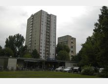 Mehrstöckige Wohngebäude aus Beton in einer städtischen Umgebung, umgeben von Bäumen. Im Vordergrund befindet sich ein niedriges Gebäude mit Fahrrädern und parkenden Autos, sowie ein Schild mit der Aufschrift „U-Bahn-Krankenhaus“.