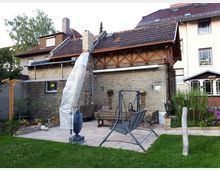 Innenhof mit gepflasterter Terrasse vor einem Backsteingebäude mit Satteldach und sichtbaren Holzstreben unter dem Dach. Auf der Terrasse stehen eine überdachte Gartenliege und Sitzbänke, umgeben von Rasen, Blumenbeeten und angrenzenden Wohnhäusern.