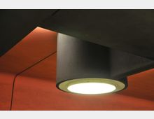 Eine Deckenleuchte mit zylindrischem Gehäuse, aus der helles Licht strömt, ist an einer Decke mit orangefarbener Beleuchtung und matter Oberfläche montiert. Die Umgebung wirkt modern und minimalistisch.