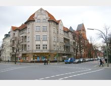 Straßenecke mit einem mehrstöckigen Wohn- und Geschäftshaus im historischen Stil, das rote Ziegeldächer und Verzierungen über den Fenstern zeigt; im Erdgeschoss befindet sich eine Apotheke. Rechts im Hintergrund ist eine große Backsteinkirche mit spitzem Turmdach zu sehen, daneben Bäume und parkende Autos.