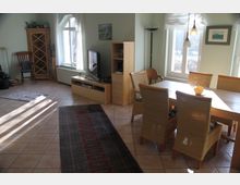 Helles Wohnzimmer mit Esstisch und sechs geflochtenen Stühlen aus Holz, Fliesenboden und Teppichen. Links befindet sich ein TV-Schrank neben einem Holzregal, und große Fenster lassen viel Tageslicht herein.