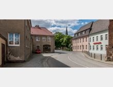 Straßenansicht einer kleinen Stadt mit gepflastertem Gehweg und einer Straße, die sich nach rechts biegt. Links stehen ältere Gebäude mit roten Dächern, rechts ein pastellfarbenes Gebäude mit der Aufschrift "Ambulanz"; im Hintergrund ist ein Kirchturm über Bäumen sichtbar.